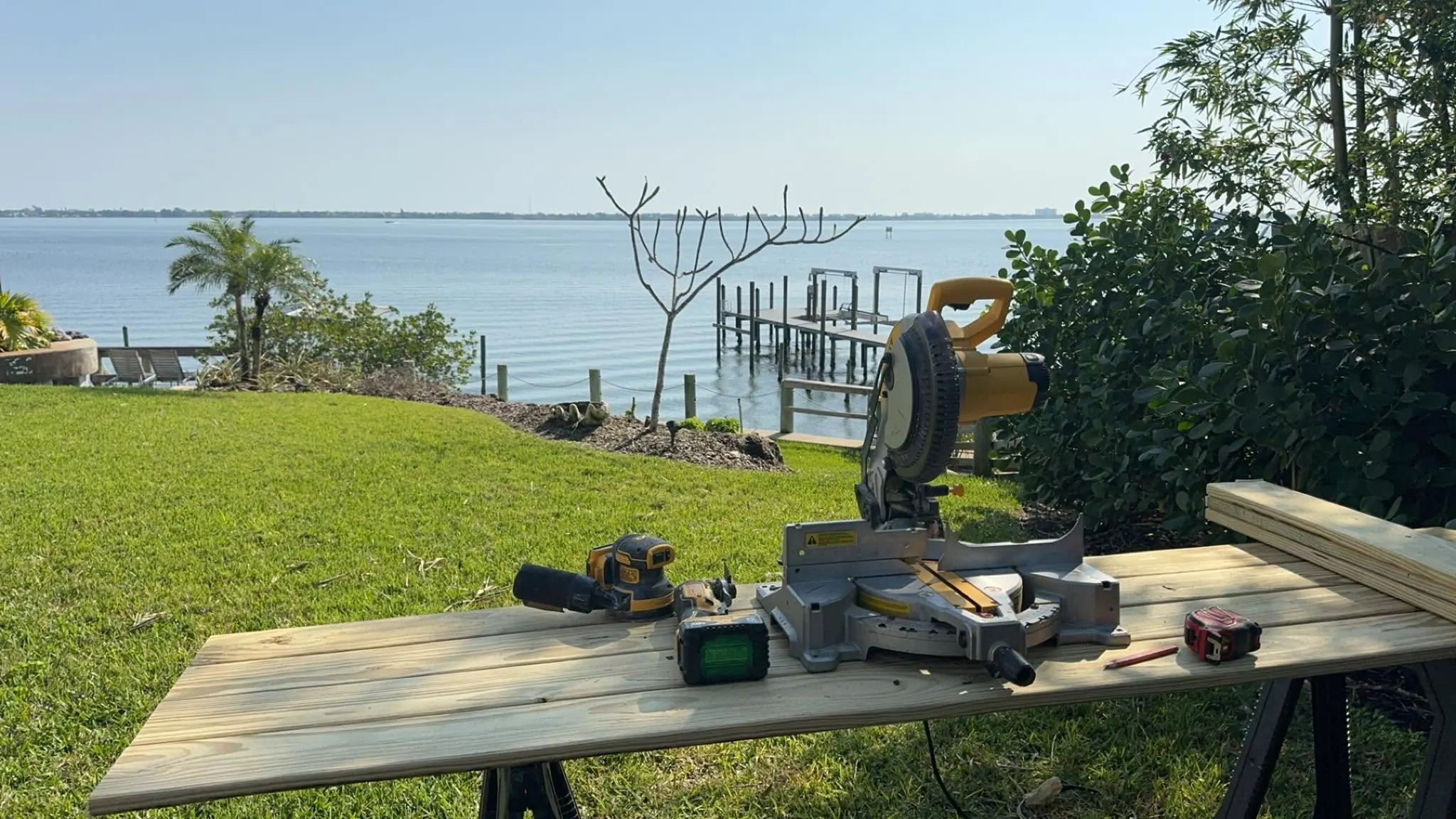 A miter saw, drill, sander and lumber set on sawhorses in a waterside yard, with a dock and a wide lagoon behind.